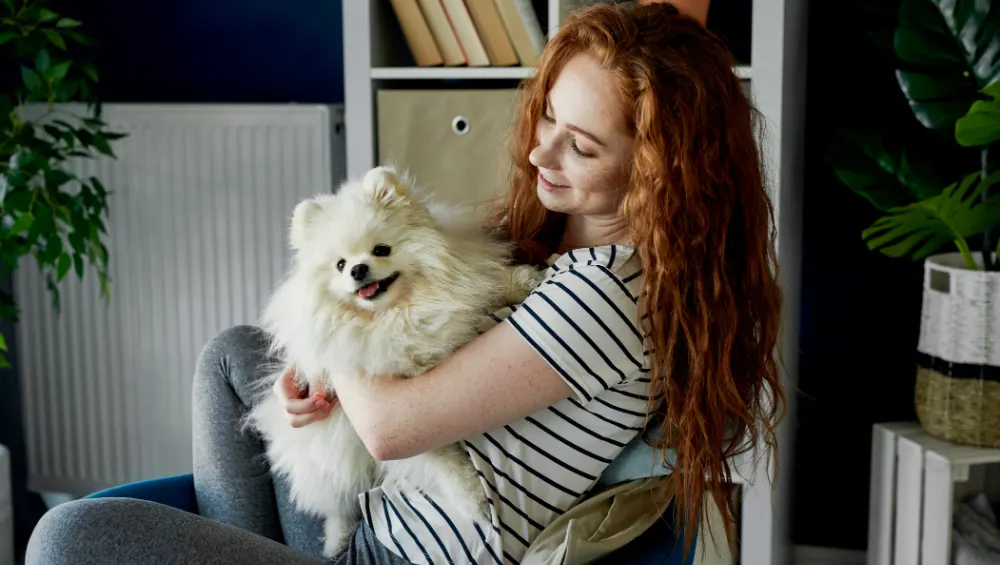A woman with long red hair holds a fluffy white Pomeranian dog while sitting in a chair.