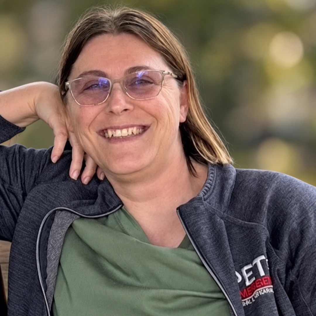 Kellyn Owens, smiling wearing glasses and a dark jacket over a green shirt, posing outdoors with one hand behind her head in a relaxed setting.