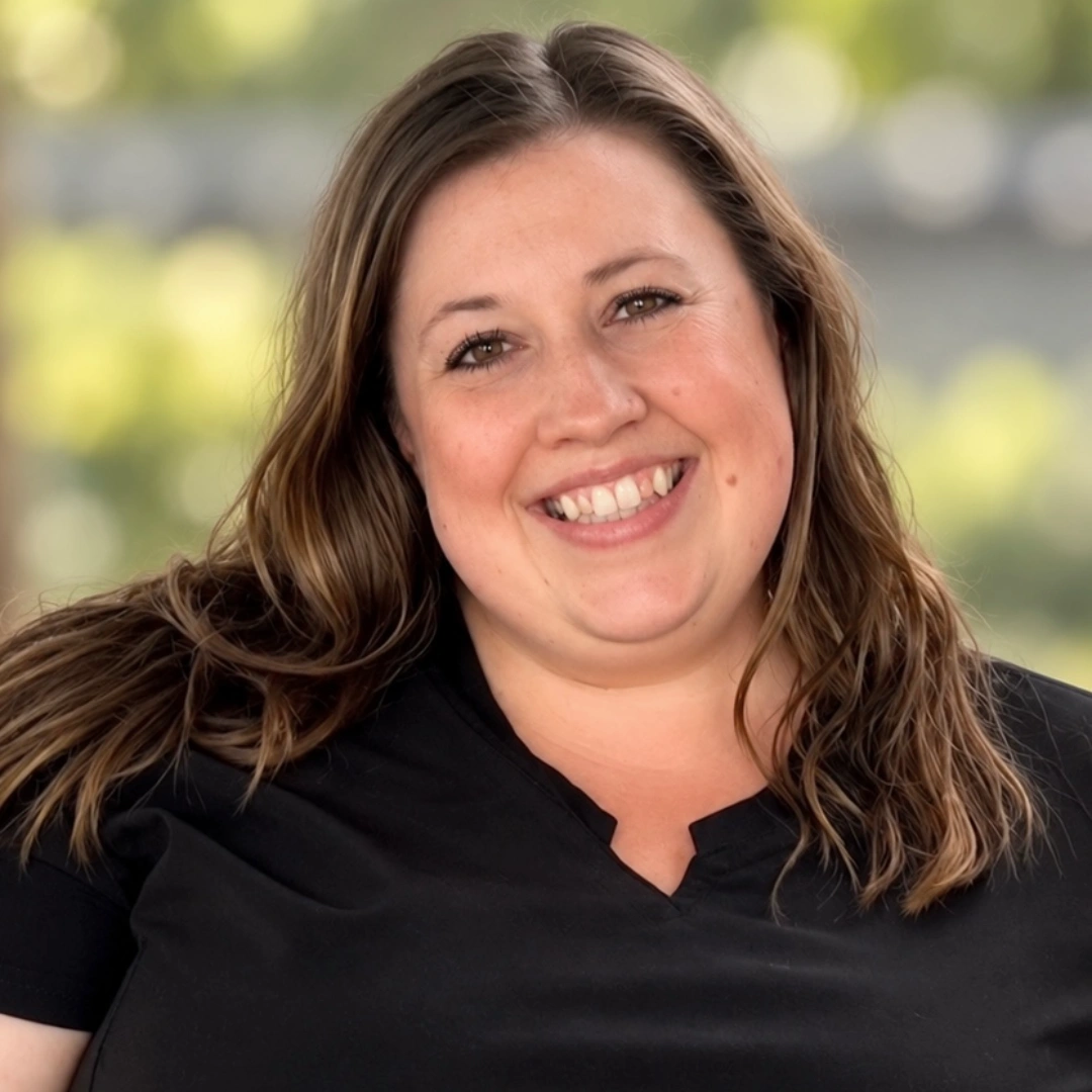 Maria Burton, smiling with long brown hair in a black top, photographed outdoors against a softly blurred green background.