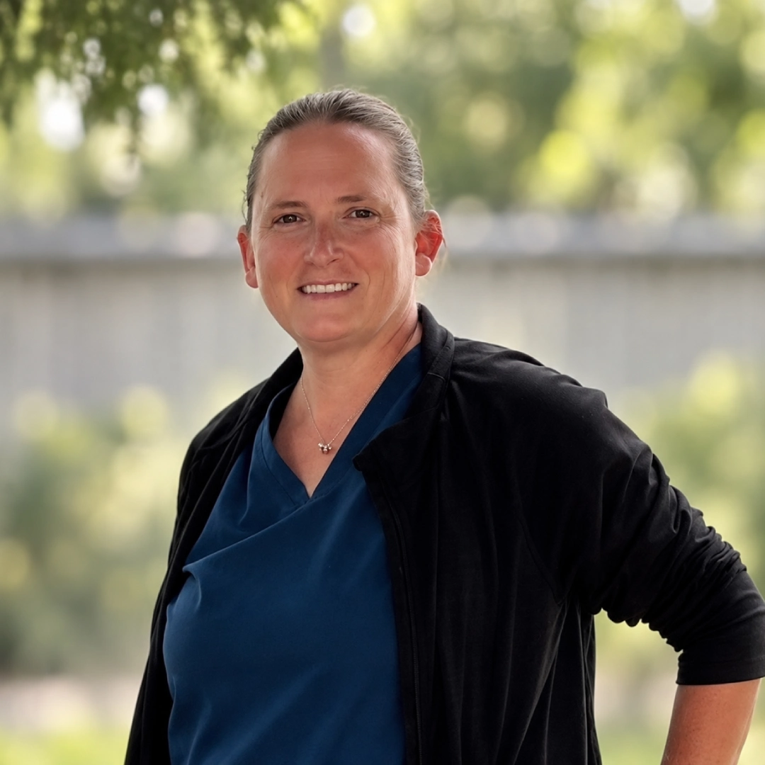 Whitney Nelson, smiling and poses outdoors in a blue blouse and black jacket, with soft green trees and a fence behind her.